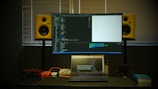 A workspace featuring a large monitor displaying code in front of a window with closed blinds. Below the monitor is a laptop open to a webpage about a developer event. On either side of the monitor are yellow studio speakers. Various items like cables, a microphone, and a book are arranged on the desk.