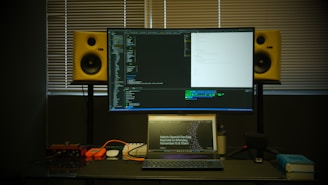A workspace featuring a large monitor displaying code in front of a window with closed blinds. Below the monitor is a laptop open to a webpage about a developer event. On either side of the monitor are yellow studio speakers. Various items like cables, a microphone, and a book are arranged on the desk.