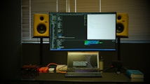 A workspace featuring a large monitor displaying code in front of a window with closed blinds. Below the monitor is a laptop open to a webpage about a developer event. On either side of the monitor are yellow studio speakers. Various items like cables, a microphone, and a book are arranged on the desk.