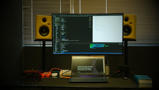 A workspace featuring a large monitor displaying code in front of a window with closed blinds. Below the monitor is a laptop open to a webpage about a developer event. On either side of the monitor are yellow studio speakers. Various items like cables, a microphone, and a book are arranged on the desk.
