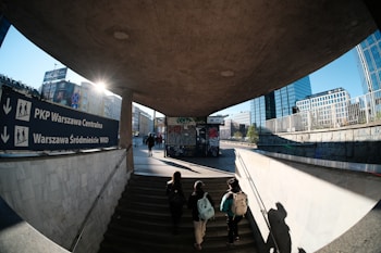 An urban scene with a group of people walking up stairs leading to a busy city street. The sun is setting or rising, casting long shadows across the ground. A large sign indicates directions to Warsaw Central and Warszawa Śr&oacute;dmieście WKD. Skyscrapers and office buildings surround the area, and graffiti is visible on nearby walls.