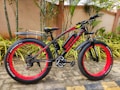 A black and red fat-tire electric bike is parked on a patio with lush greenery and palm plants in the background. It features the brand name 'Voltbik' on the frame and tires, the bike has a sturdy frame and wide handlebars. The ground is paved with stone tiles, and the background wall is a muted beige color.