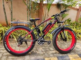 A black and red fat-tire electric bike is parked on a patio with lush greenery and palm plants in the background. It features the brand name 'Voltbik' on the frame and tires, the bike has a sturdy frame and wide handlebars. The ground is paved with stone tiles, and the background wall is a muted beige color.