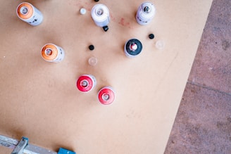 Several spray paint cans of different colors, including red, orange, and black, are scattered on a wooden surface. Some caps are off, and a few are set aside. The surface shows signs of paint marks.