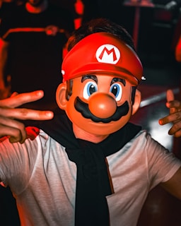 A person is wearing a large plastic Mario mask with exaggerated features, including a big nose and mustache. They are dressed casually in a white shirt and have a black garment draped around their shoulders. The background is dimly lit with red lighting, suggesting a festive or party atmosphere. The individual is giving a peace sign with their fingers.