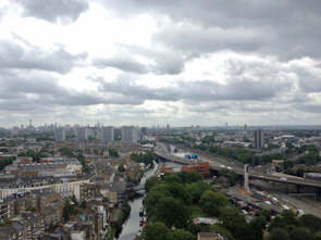 An urban landscape featuring a city skyline with prominent tall buildings under a cloudy sky. In the foreground, a river meanders through a residential area with a mixture of houses and green spaces. Roads and railways are visible, suggesting a busy, interconnected city.