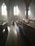 Sunlight streams through the tall, arched windows of an empty church, casting long shadows across the wooden pews and creating an ethereal glow. The atmosphere is calm and serene, enhanced by the soft haze of light.