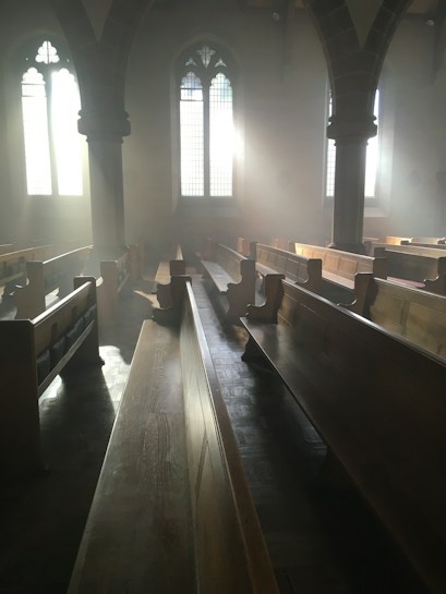 Sunlight streams through the tall, arched windows of an empty church, casting long shadows across the wooden pews and creating an ethereal glow. The atmosphere is calm and serene, enhanced by the soft haze of light.