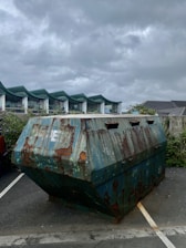A rusty, weathered metal dumpster is situated in a parking area. The sky overhead is overcast with grey clouds. Behind the dumpster, there are buildings with modern architecture featuring green-tinted roof structures. There are also some plants and vegetation visible near the buildings.