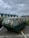 A rusty, weathered metal dumpster is situated in a parking area. The sky overhead is overcast with grey clouds. Behind the dumpster, there are buildings with modern architecture featuring green-tinted roof structures. There are also some plants and vegetation visible near the buildings.