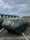 A rusty, weathered metal dumpster is situated in a parking area. The sky overhead is overcast with grey clouds. Behind the dumpster, there are buildings with modern architecture featuring green-tinted roof structures. There are also some plants and vegetation visible near the buildings.
