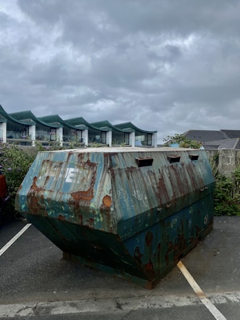 A rusty, weathered metal dumpster is situated in a parking area. The sky overhead is overcast with grey clouds. Behind the dumpster, there are buildings with modern architecture featuring green-tinted roof structures. There are also some plants and vegetation visible near the buildings.