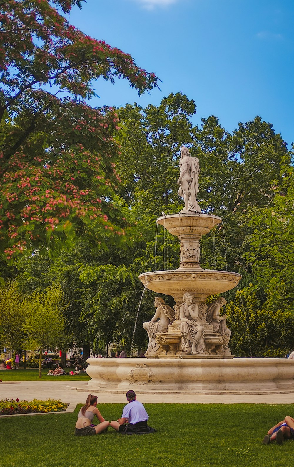 two people sitting on the grass near a fountain