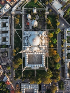 An aerial view of a large mosque complex with multiple domes and courtyards, surrounded by green gardens and trees. The architecture is detailed with symmetrical designs and tall minarets. The surrounding area includes urban buildings and roads.