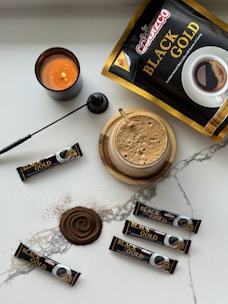 A neatly arranged setup featuring a candle, a large packet of 'Black Gold' coffee, several smaller sticks of instant coffee, a cup of frothy coffee, a black spoon, and a swirl of coffee powder on a light marble surface.