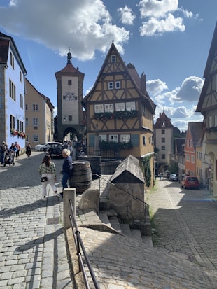A picturesque medieval street scene with half-timbered buildings, cobblestone paths, and a stone tower with an ornate clock. People stroll on the street, and the sun casts playful shadows, with fluffy clouds in the bright blue sky.
