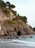 A rugged cliffside adorned with lush vegetation and a building labeled 'Hotel Porto Roca Ristorante' perched near the top. The rocky cliff drops down to a beach with gentle turquoise waves washing over the stones. Trees and shrubs grow along the cliffside, adding a verdant touch to the landscape.