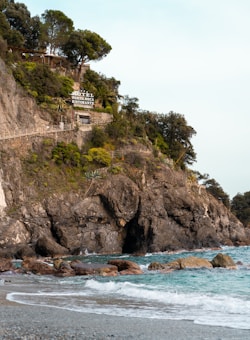 A rugged cliffside adorned with lush vegetation and a building labeled 'Hotel Porto Roca Ristorante' perched near the top. The rocky cliff drops down to a beach with gentle turquoise waves washing over the stones. Trees and shrubs grow along the cliffside, adding a verdant touch to the landscape.