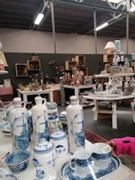 A collection of blue and white ceramic items, including bottles, bowls, and teapots, is displayed on a table. The ceramics feature intricate patterns and depictions of windmills. Surrounding these are various decorative objects like glass jars, figurines, and lamps arranged on shelves and tables in an antique shop setting.
