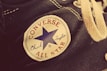 A close-up view of a Converse All Star Chuck Taylor canvas shoe, highlighting the iconic patch with a blue star, the words Converse All Star in red, and Chuck Taylor in blue script. The shoe features textured fabric and cream-colored laces with metal eyelets.
