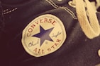 A close-up view of a Converse All Star Chuck Taylor canvas shoe, highlighting the iconic patch with a blue star, the words Converse All Star in red, and Chuck Taylor in blue script. The shoe features textured fabric and cream-colored laces with metal eyelets.