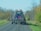 A large blue agricultural implement being towed on a rural road. The machinery has a series of metal prongs or discs and is folded up for transport. The road is surrounded by trees with sparse leaves, indicating a possible late autumn setting.