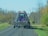 A large blue agricultural implement being towed on a rural road. The machinery has a series of metal prongs or discs and is folded up for transport. The road is surrounded by trees with sparse leaves, indicating a possible late autumn setting.