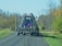 A large blue agricultural implement being towed on a rural road. The machinery has a series of metal prongs or discs and is folded up for transport. The road is surrounded by trees with sparse leaves, indicating a possible late autumn setting.