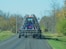 A large blue agricultural implement being towed on a rural road. The machinery has a series of metal prongs or discs and is folded up for transport. The road is surrounded by trees with sparse leaves, indicating a possible late autumn setting.