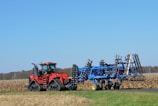 A large red tractor with tracks instead of wheels is pulling a blue agricultural implement through a vast cornfield. The sky is clear and blue, indicating a sunny day. The corn stalks are dry and golden, suggesting the harvest season.