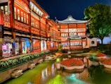 A traditional Chinese building with intricate architectural details illuminated by warm lights. A serene garden pond with floating flower arrangements reflects the building's vibrant colors. Lush greenery and a large tree enhance the tranquil atmosphere.