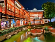 A traditional Chinese building with intricate architectural details illuminated by warm lights. A serene garden pond with floating flower arrangements reflects the building's vibrant colors. Lush greenery and a large tree enhance the tranquil atmosphere.