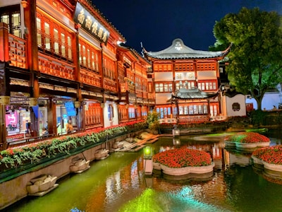 A traditional Chinese building with intricate architectural details illuminated by warm lights. A serene garden pond with floating flower arrangements reflects the building's vibrant colors. Lush greenery and a large tree enhance the tranquil atmosphere.