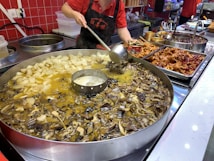 A person in a red shirt and black apron is cooking using a large circular pan filled with various types of food, including mushrooms and sliced vegetables, immersed in a broth. There are additional prepared dishes and ingredients on the countertop nearby, including spicy items and some chicken parts.