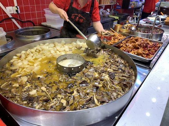 A person in a red shirt and black apron is cooking using a large circular pan filled with various types of food, including mushrooms and sliced vegetables, immersed in a broth. There are additional prepared dishes and ingredients on the countertop nearby, including spicy items and some chicken parts.