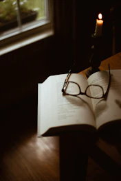 An open book resting on a wooden table with a pair of reading glasses and a lit candle nearby.