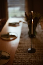 A dimly lit dining table with a textured table runner is set with plates and napkins. A single lit candle on a candle holder sits on the table, casting a warm glow. A vase of flowers is partially visible in the background. The atmosphere is serene and elegant.