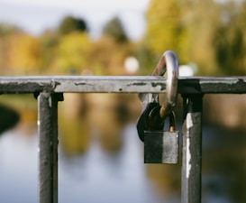 A padlock is secured on a metal railing with a blurred natural background. The railing shows signs of wear and chipped paint, while the out-of-focus backdrop suggests greenery and possibly a body of water.