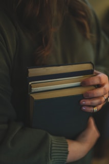 A person wearing a dark green sweater is holding several hardcover books with one arm, pressing them gently against their body. The person's fingers are adorned with rings, and their nails are painted. The scene is softly lit, creating a warm and intimate atmosphere.