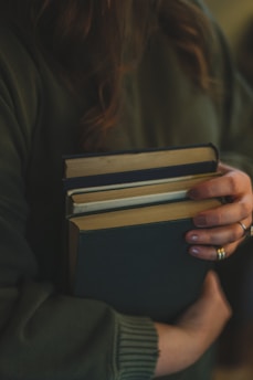 A person wearing a dark green sweater is holding several hardcover books with one arm, pressing them gently against their body. The person's fingers are adorned with rings, and their nails are painted. The scene is softly lit, creating a warm and intimate atmosphere.