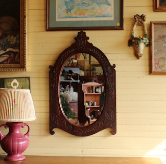 An ornate, oval-shaped mirror with a wooden frame is mounted on a yellow wooden wall. The wall is decorated with various framed artworks and a floral wall sconce. A pink ceramic lamp with a fabric lampshade sits on a wooden table. Inside the mirror's reflection, shelves with various objects, including colorful dishes and decorative items, are visible.
