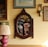 An ornate, oval-shaped mirror with a wooden frame is mounted on a yellow wooden wall. The wall is decorated with various framed artworks and a floral wall sconce. A pink ceramic lamp with a fabric lampshade sits on a wooden table. Inside the mirror's reflection, shelves with various objects, including colorful dishes and decorative items, are visible.