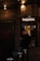 A person is standing in front of a ticket booth labeled 'TAQUILLA' with a light overhead. The setting is dimly lit, with brick walls and string lights providing a warm ambiance. The person appears to be handling a phone or device.