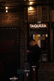 A person is standing in front of a ticket booth labeled 'TAQUILLA' with a light overhead. The setting is dimly lit, with brick walls and string lights providing a warm ambiance. The person appears to be handling a phone or device.