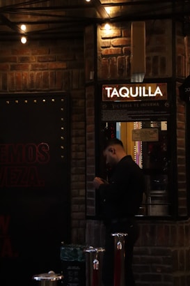 A person is standing in front of a ticket booth labeled 'TAQUILLA' with a light overhead. The setting is dimly lit, with brick walls and string lights providing a warm ambiance. The person appears to be handling a phone or device.