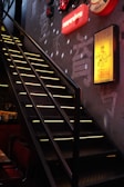 A dimly lit staircase with yellow light strips illuminating the steps, leading upwards against a textured wall. Neon signs with various characters and designs adorn the wall, adding a colorful glow to the space. The ambiance suggests an urban or modern setting, likely in a bar or restaurant.