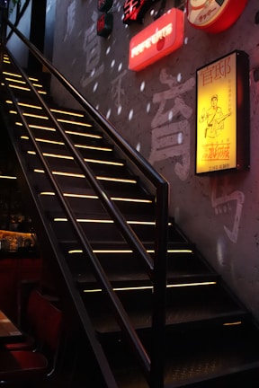 A dimly lit staircase with yellow light strips illuminating the steps, leading upwards against a textured wall. Neon signs with various characters and designs adorn the wall, adding a colorful glow to the space. The ambiance suggests an urban or modern setting, likely in a bar or restaurant.