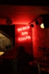 A dimly lit interior with a neon sign saying 'Beber Sin Culpa' on a wooden-paneled wall. A person is seated at a table in the foreground, creating a cozy, intimate atmosphere. The red and warm tones from the neon sign dominate, casting a soft glow over the scene.