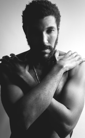 A monochromatic photograph features a bearded man with his arms crossed over his bare chest, standing against a plain background. The lighting is dramatic with shadows emphasizing the contours of his face and body.
