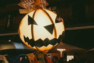 A large, illuminated jack-o'-lantern with a menacing smile and triangular eyes dominates the foreground. It appears to be part of a Halloween celebration, with the word 'HALLOWEEN' visible on a sign above. The background is dark and suggests a nighttime setting, possibly with people and decorations present.